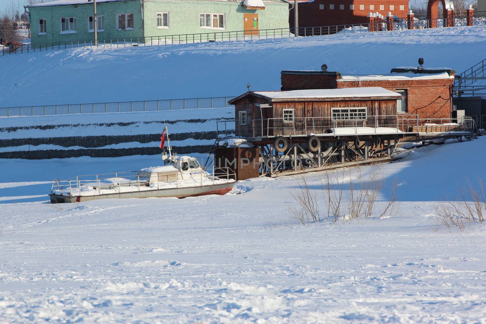 The frozen river port. Cranes, barges, old buildings. An ice-covered river covered with snow and a dry forest in the background