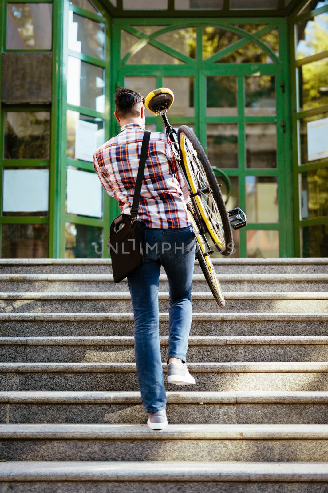 Casual businessman going to work by bicycle. He is carrying the bike upstairs to the entrance of the office building. Rear view.