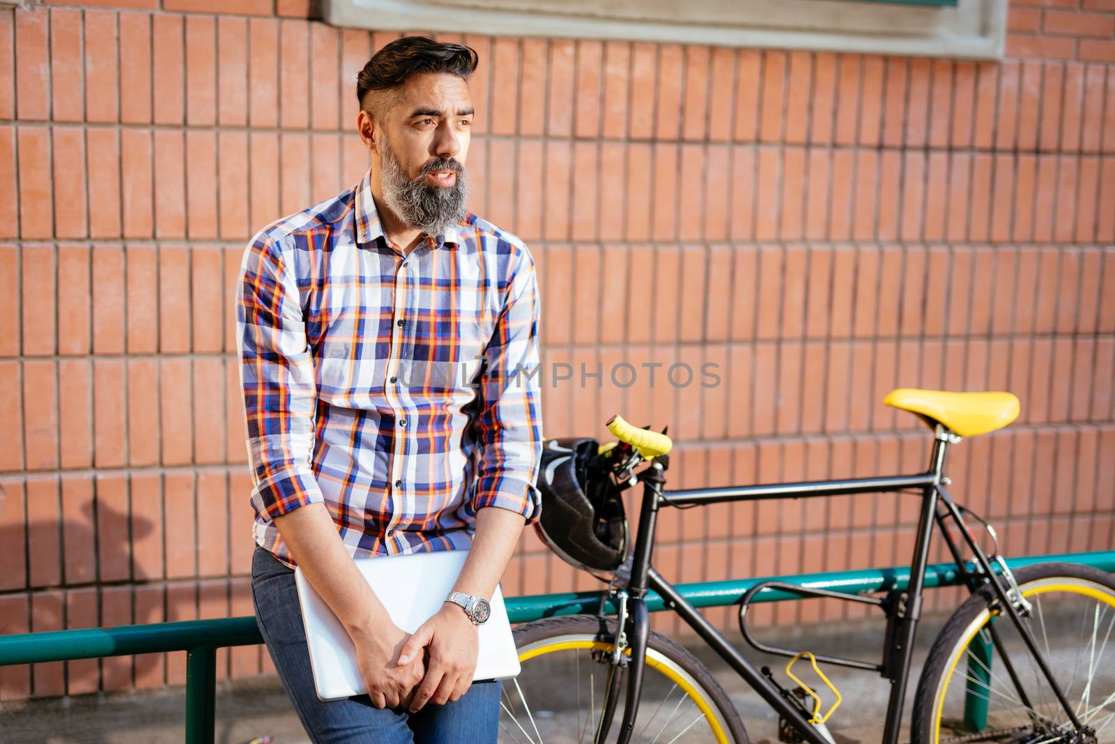 Casual businessman having a break. He is standing in front of the building next to bike and holding laptop.