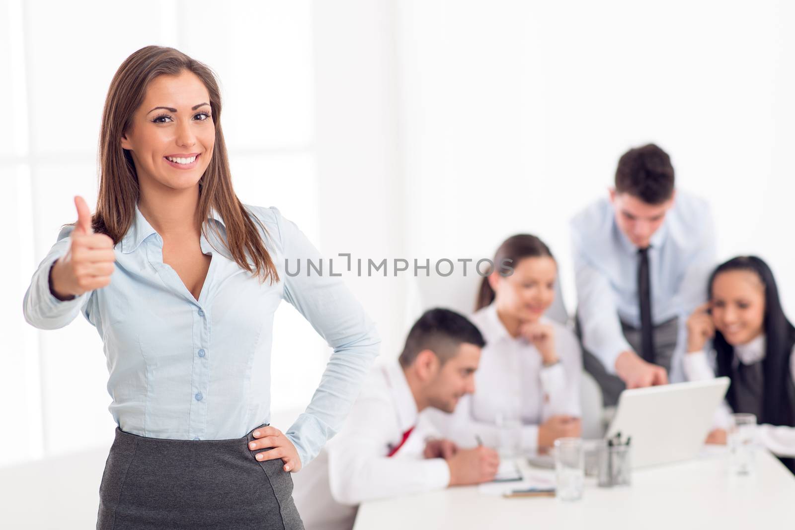 Successful young businesswoman standing proudly in the office and looking at the camera. Her young colleagues working at laptop on desk.