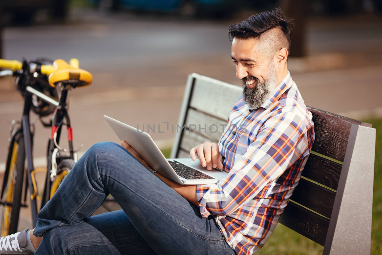Smiling casual businessman on a break. He is sitting on a bench and working at laptop, next the bench rests a bike.