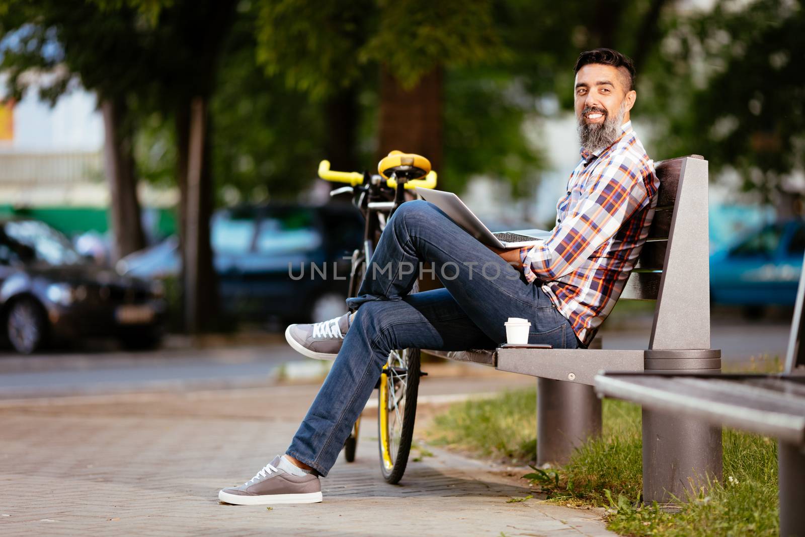 Casual businessman on a coffee break. He is sitting on a bench and working at laptop, next the bench rests a bike.