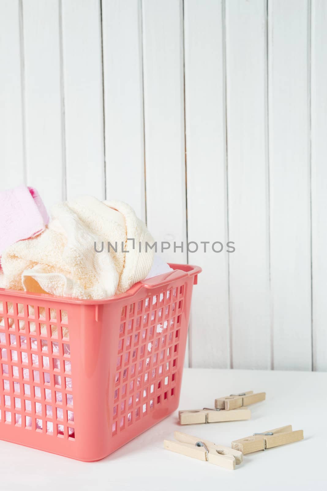 White, beige and pink towels in a red plastic laundry basket and wooden clothespins are on the white wooden background