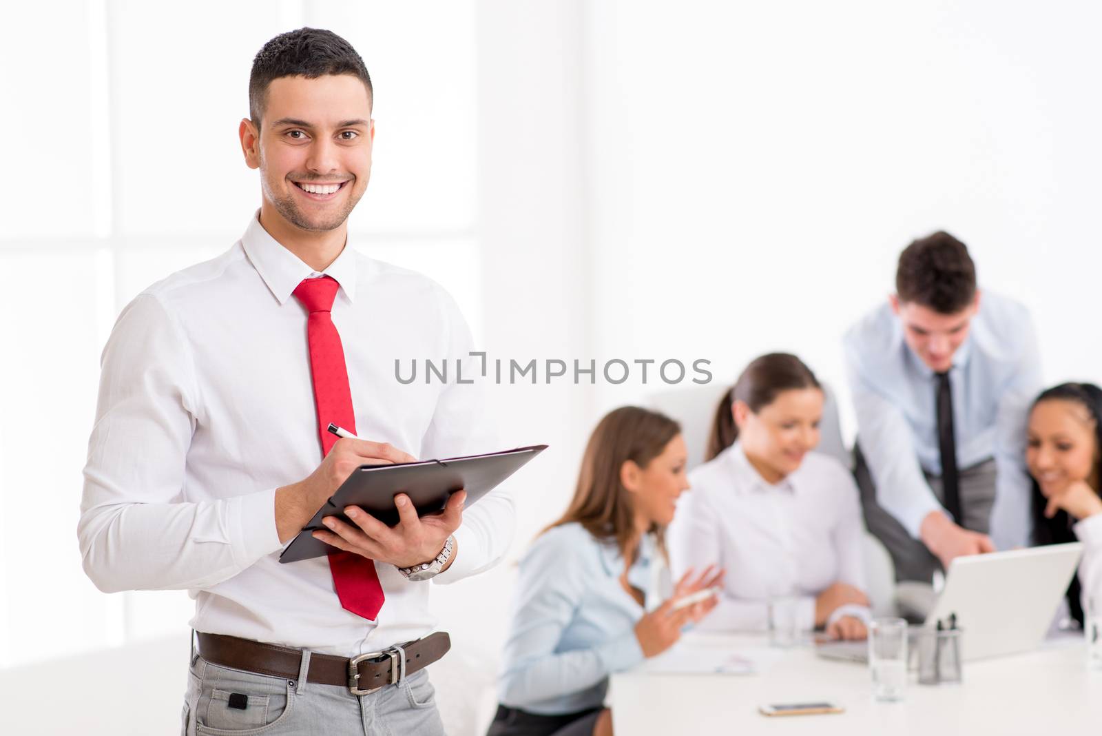 Successful young businessman standing proudly in the office and looking at the camera. Her young colleagues working at laptop on desk.