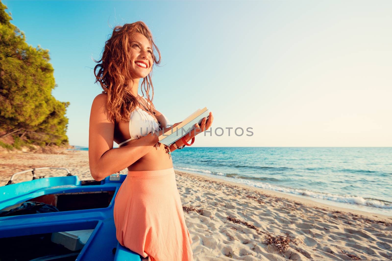 Young woman reading a book on the sandy beach.  She is standing next to boat and looking away with smile on her thinking face.