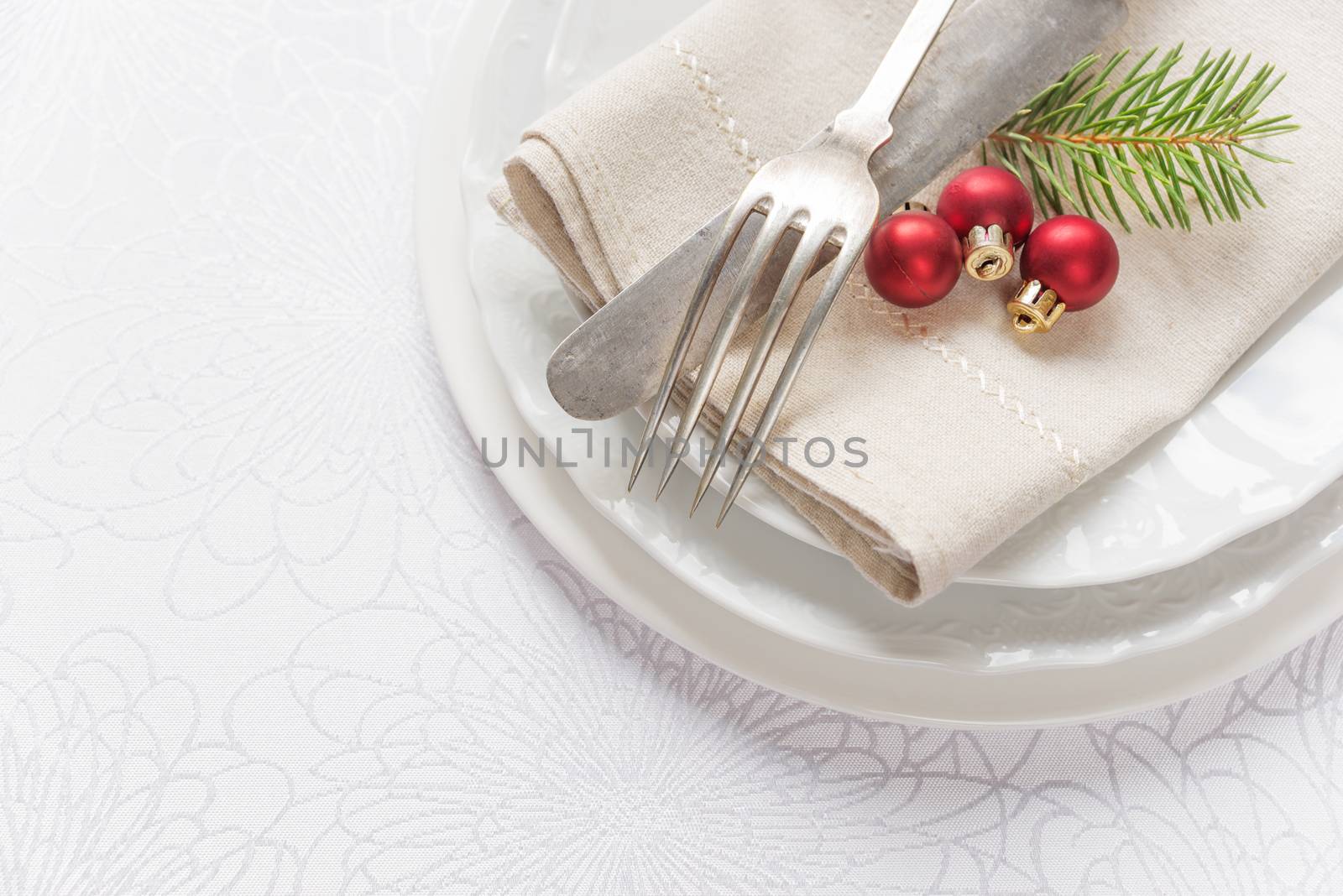 Old knife and fork, three red christmas balls and green spruce branche lie on the white porcelain plate, which is located on a table covered with a white tablecloth