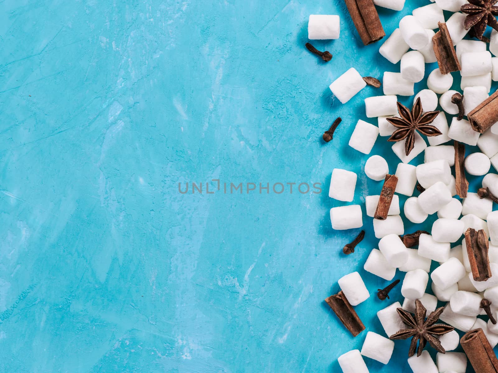 Marshmallows and winter spices on blue background with copyspace. Flat lay or top view. Background of colorful mini marshmallows, cinnamon, cloves and star anise. Winter food background concept.