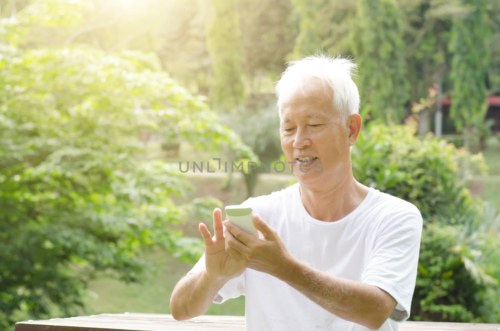 Portrait of cheerful gray hair Asian old man using smart phone, sitting at outdoor park in morning. 