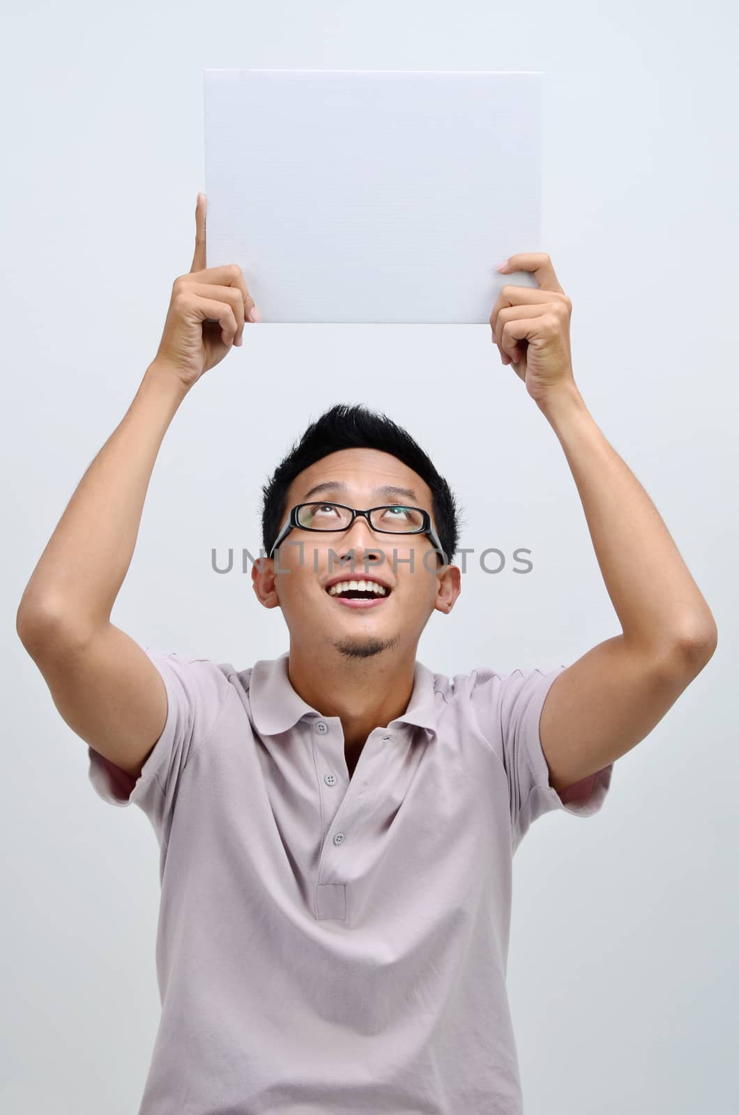 Portrait of excited Asian man hand holding white blank paper card above his head, standing isolated on plain background.