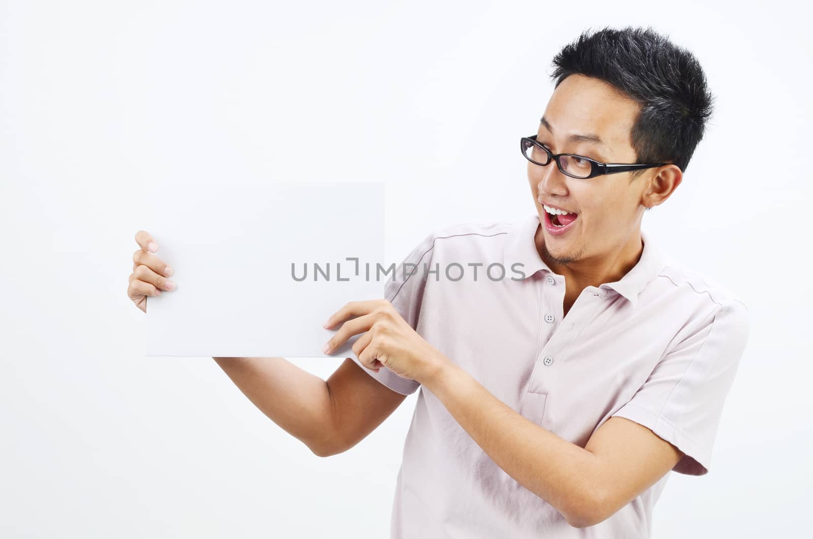 Portrait of surprised Asian man hand holding white blank paper card, standing isolated on plain background.