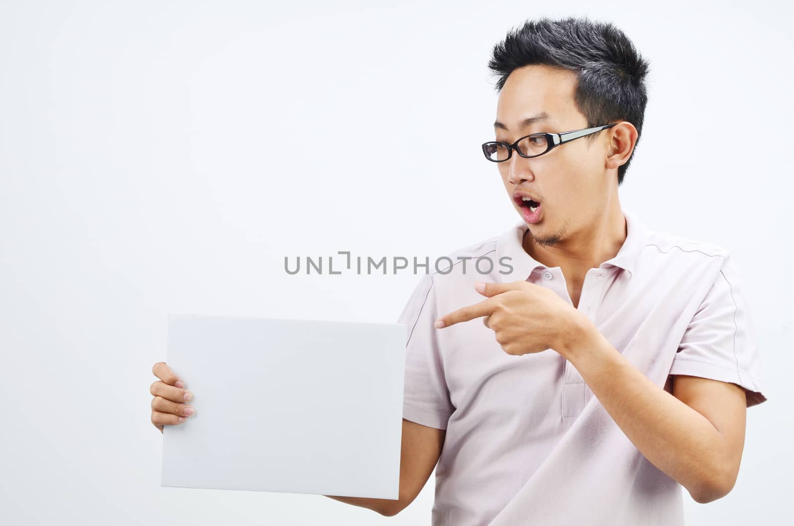 Portrait of shocked Asian man hand holding white blank paper card, standing isolated on plain background.