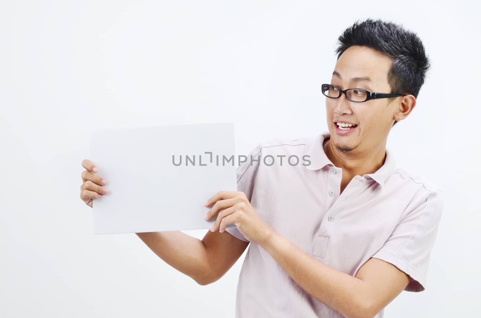 Portrait of excited Asian man hand holding white blank paper card, standing isolated on plain background.