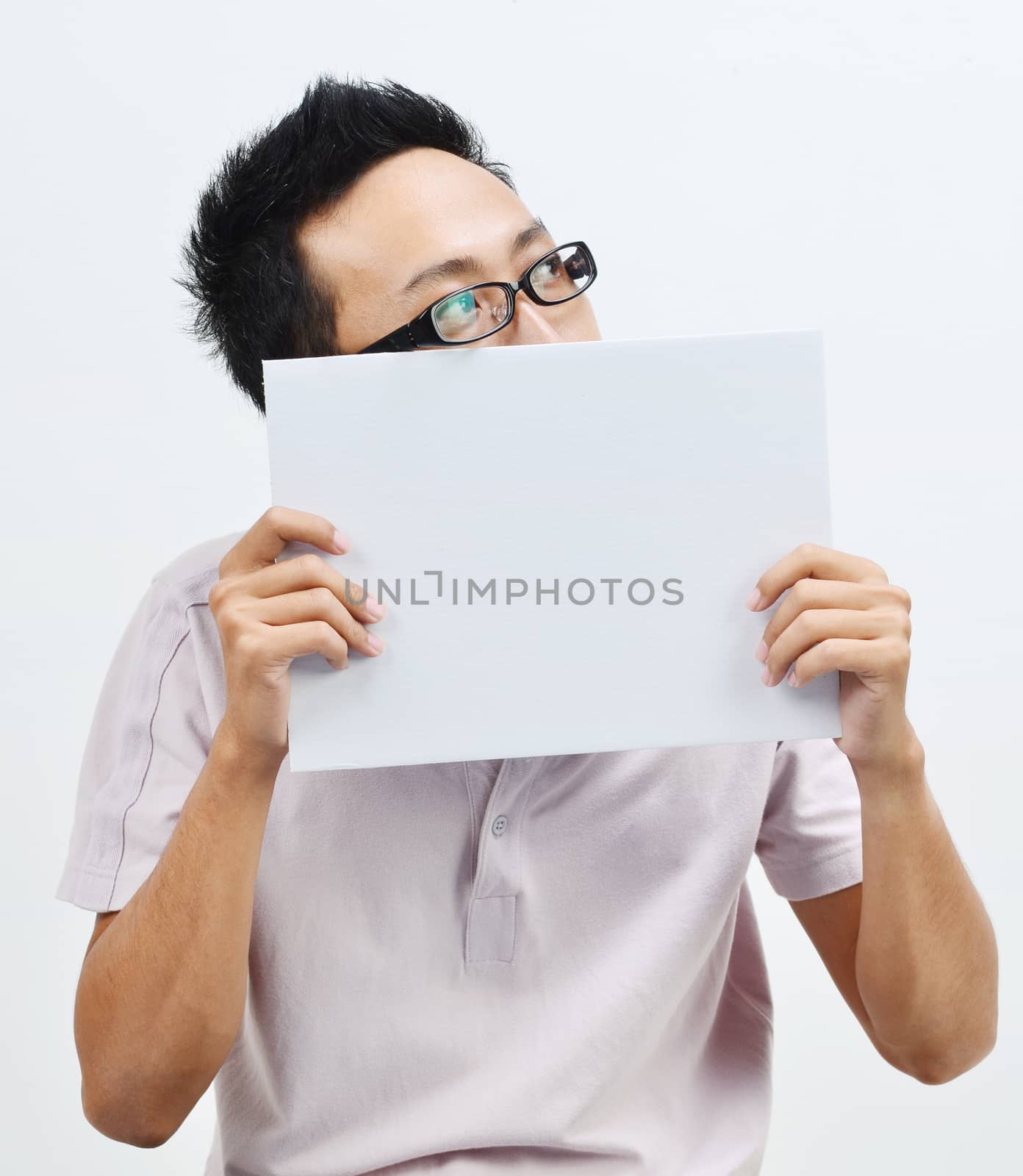 Portrait of Asian man hand holding white blank paper card covering his mouth and looking at side, standing isolated on plain background.