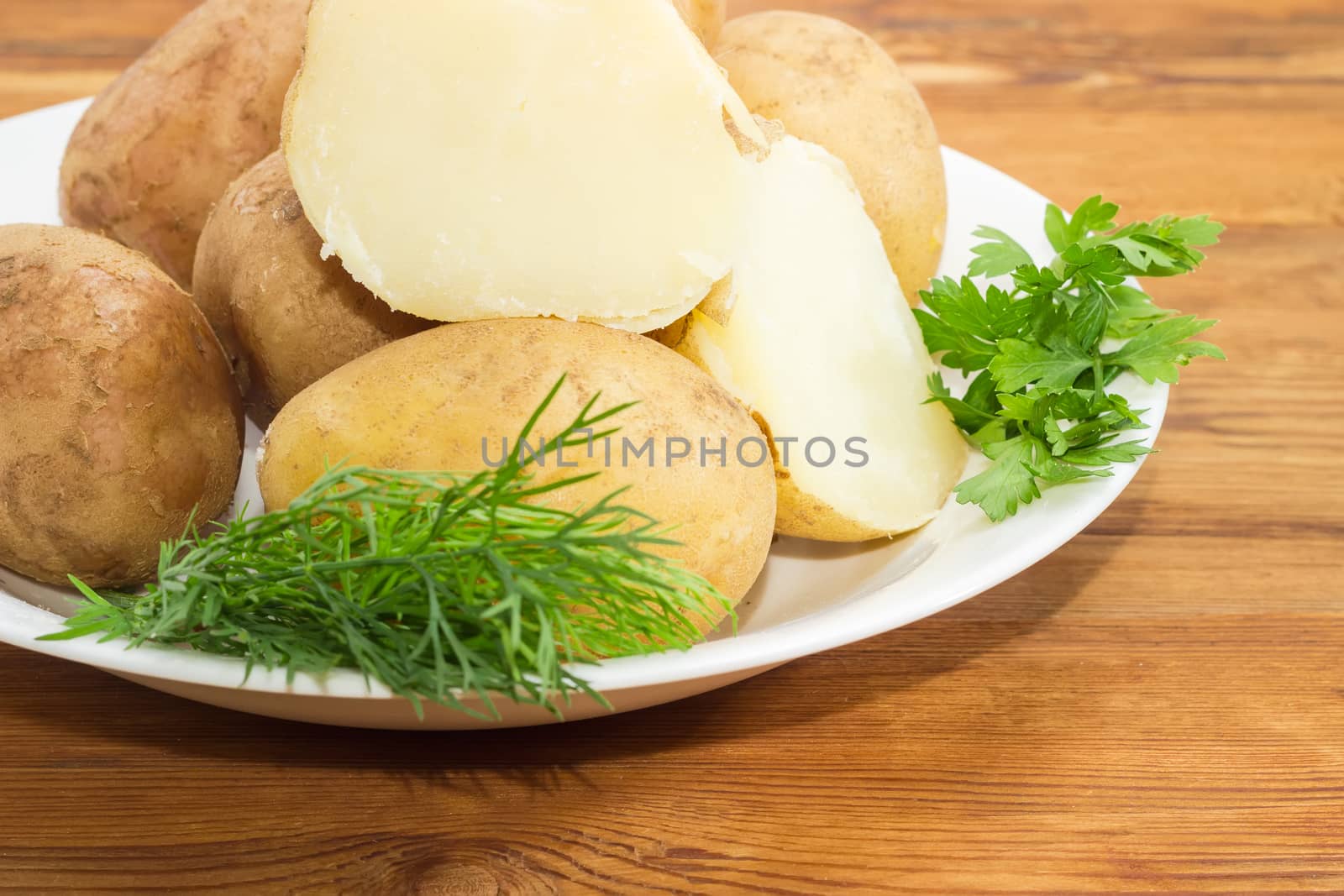 Fragment of white dish with potatoes boiled in their jackets decorated with twigs of parsley and dill on a surface of old wooden planks closeup
