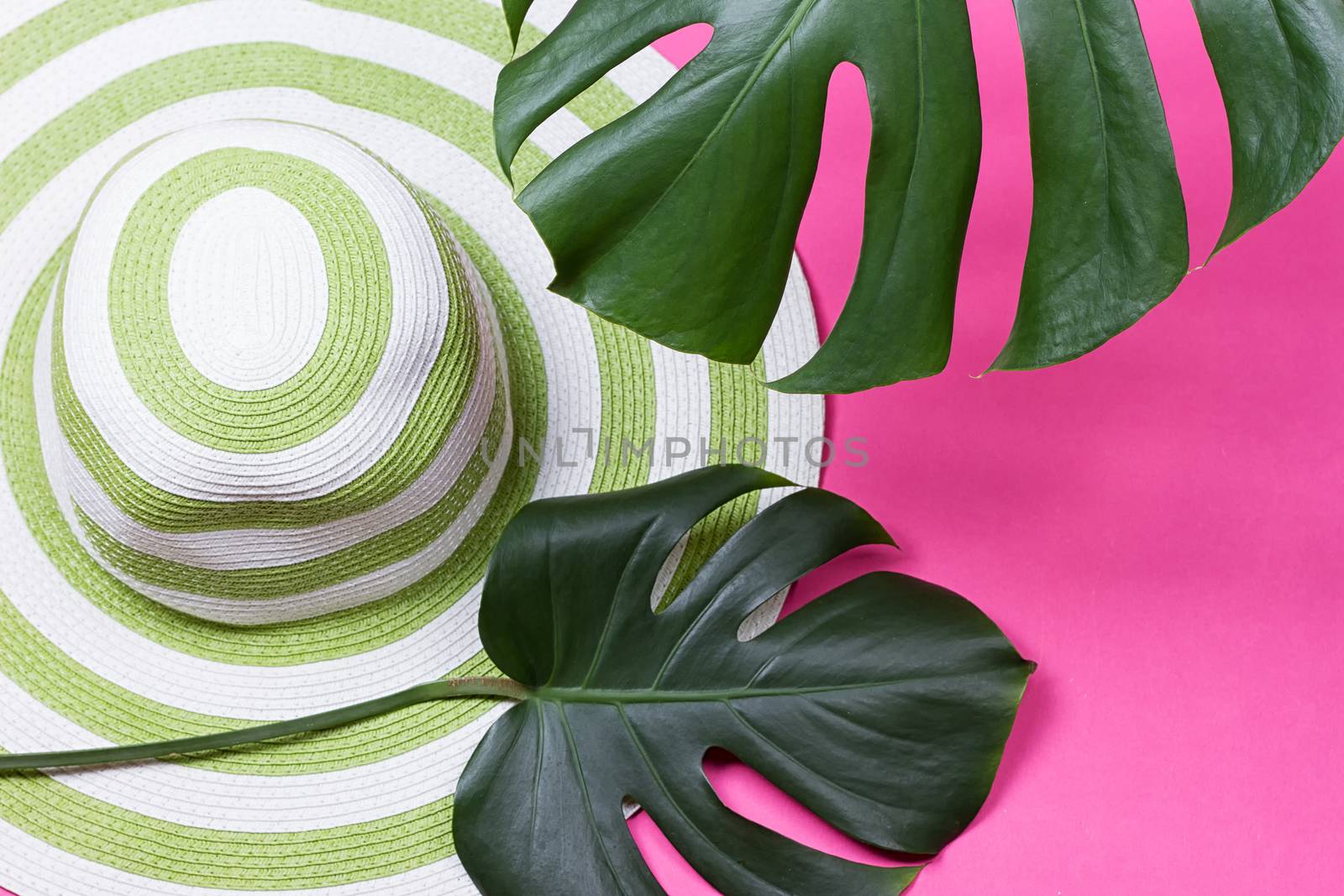 on green monstera leaf with sunglasses and straw hat on pink marble table