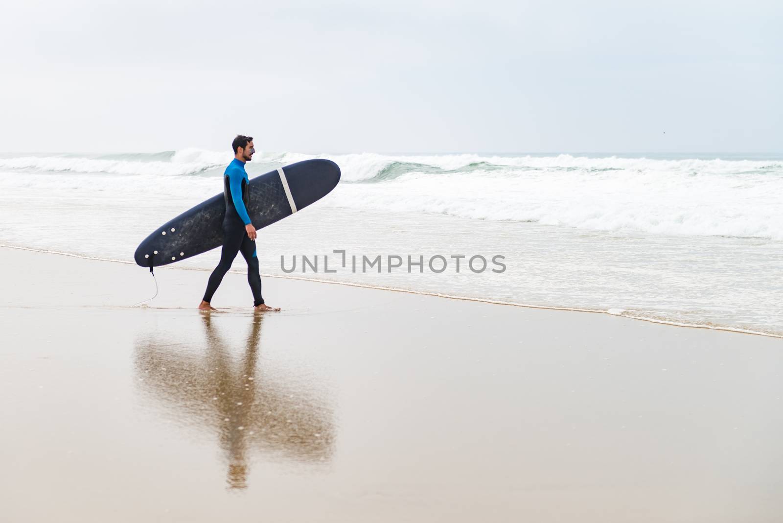 Young male surfer wearing wetsuit, holding surfboard under his arm, walking on beach after morning surfing session.