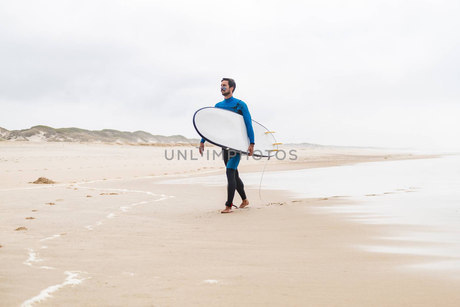 Young male surfer wearing wetsuit, holding surfboard under his arm, walking on beach after morning surfing session.