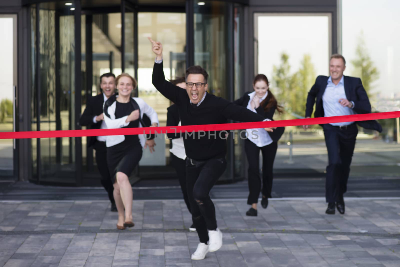 Group of happy business people running from office building crossing red ribbon finish line