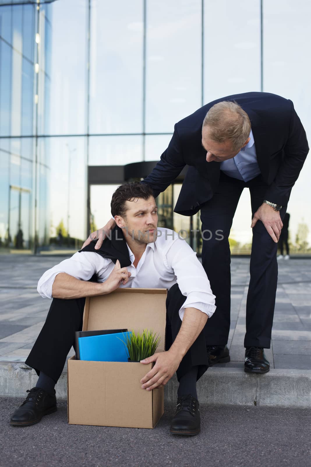 Fired business man sitting frustrated and upset on the street near office building with box of his belongings. He lost work. Other businessman comforts and encourages him