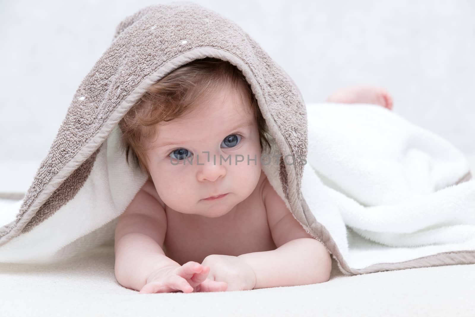 Cute newborn baby girl with beautiful blue eyes on a white terry coverlet. Adorable baby looking out under a white blanket or towel. Sad baby girl in white towel after shower