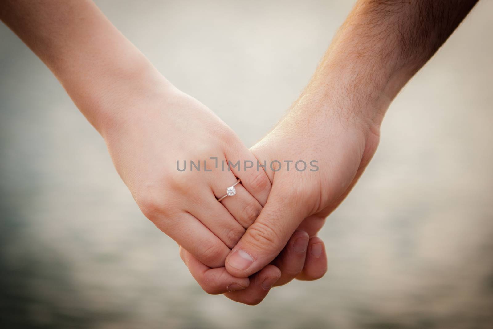 Hands holding on a beautiful blurred water background. One hand has an engagement ring.