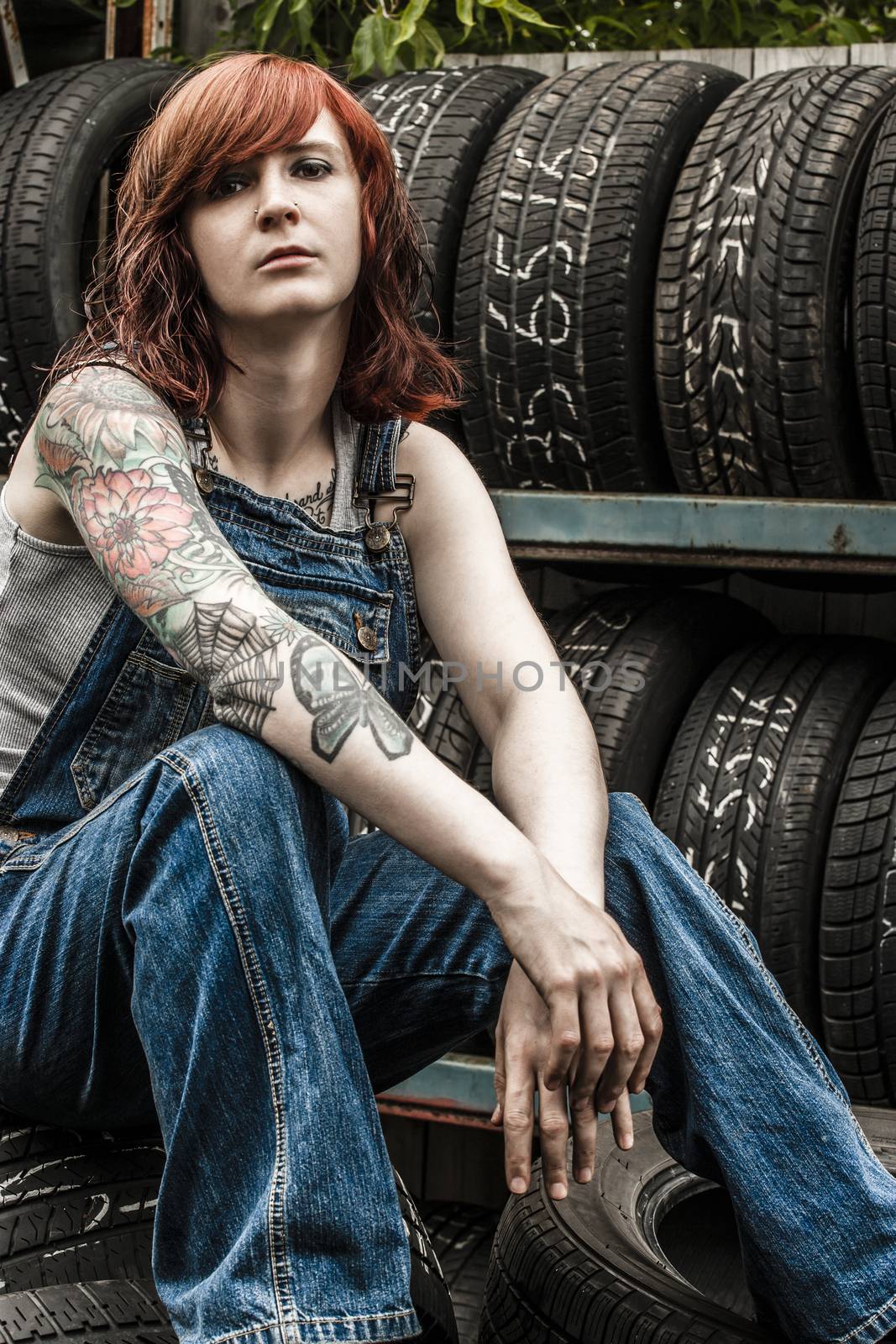 Photo of a young beautiful redhead mechanic wearing overalls and sitting behind an old garage. Attached property release is for arm tattoos.