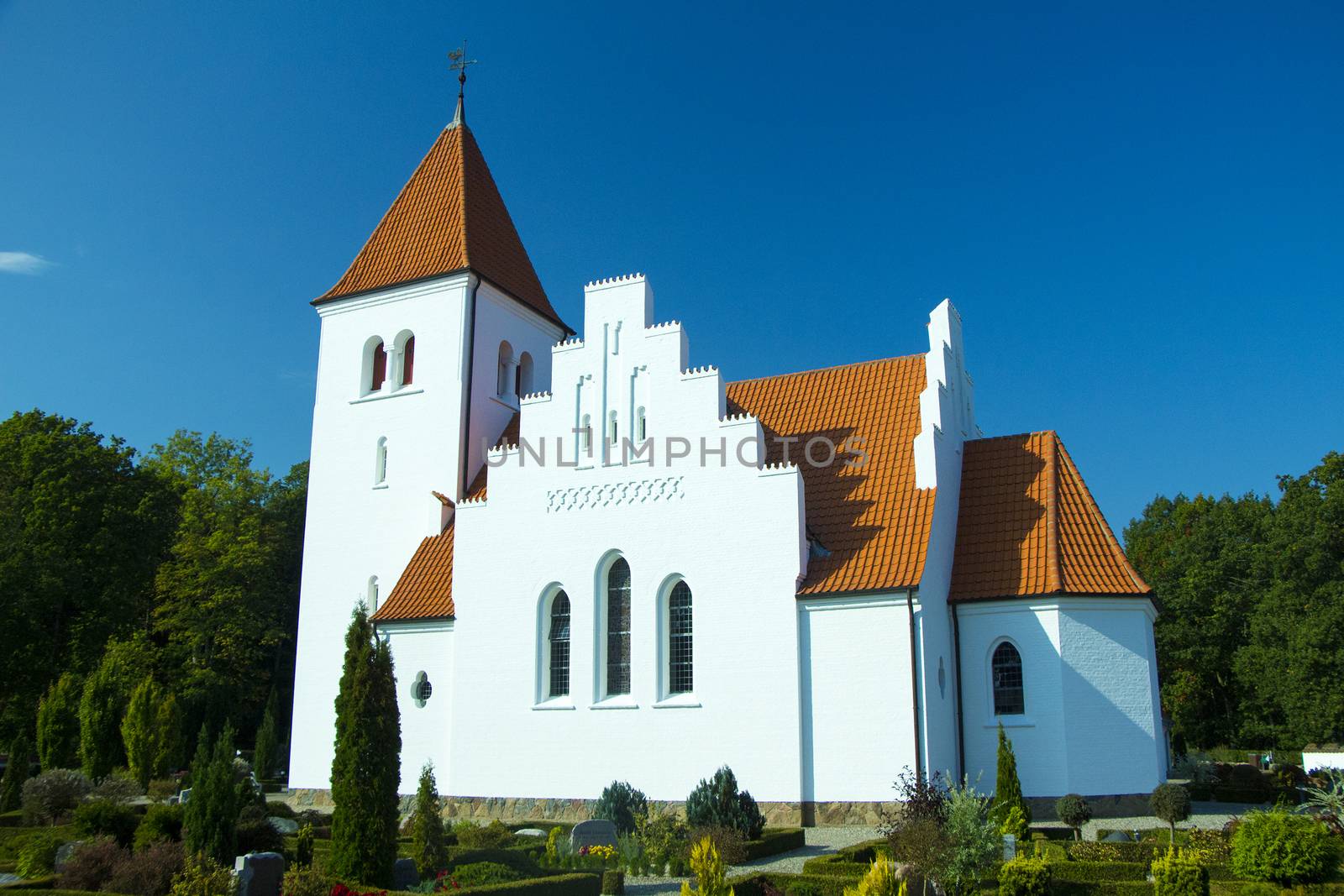 White church with orange tiles surrounded by graveyard with plants and a blue sky on a sunny day.