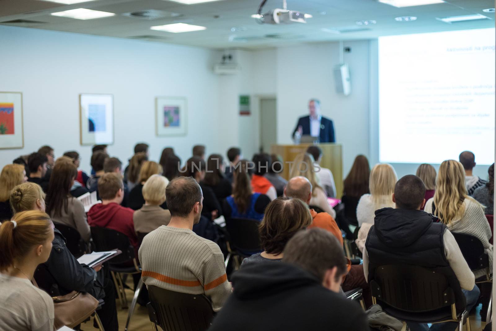 Male speaker giving presentation in lecture hall at university workshop. Audience in conference hall. Rear view of unrecognized participant in audience. Scientific conference event.