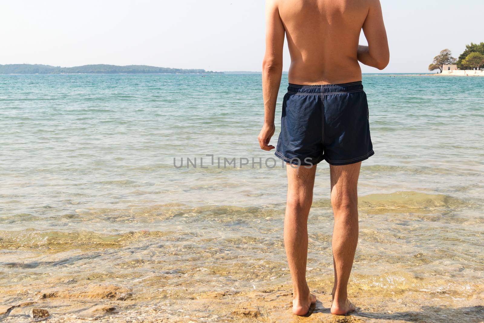 Masculine model of backs looking at the sea. Clear water, bright sun and sunscreen for sunburn. Man in blue shorts getting ready to go swimming in the mediterranean sea