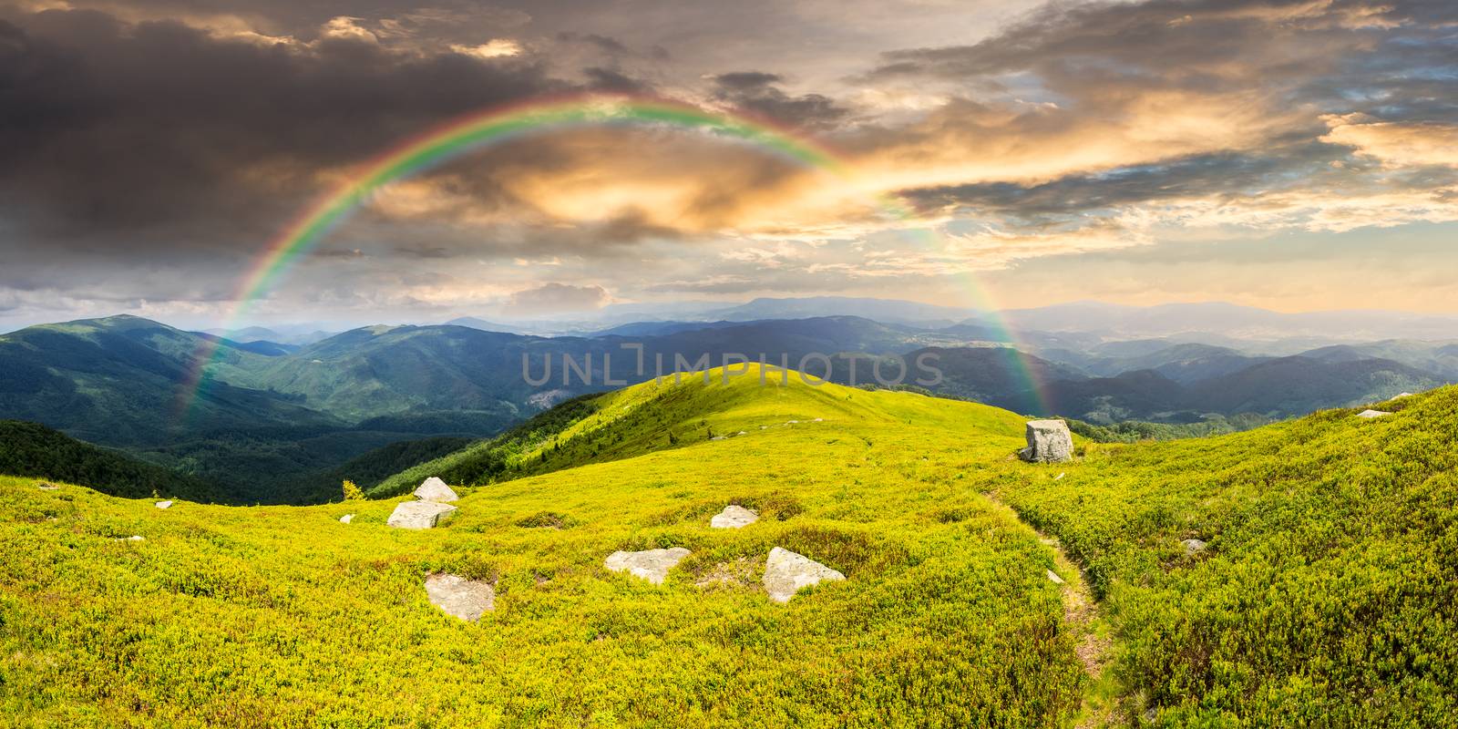 composite landscape with narrow meadow path in grass among white stones on top of mountain range with rainbow