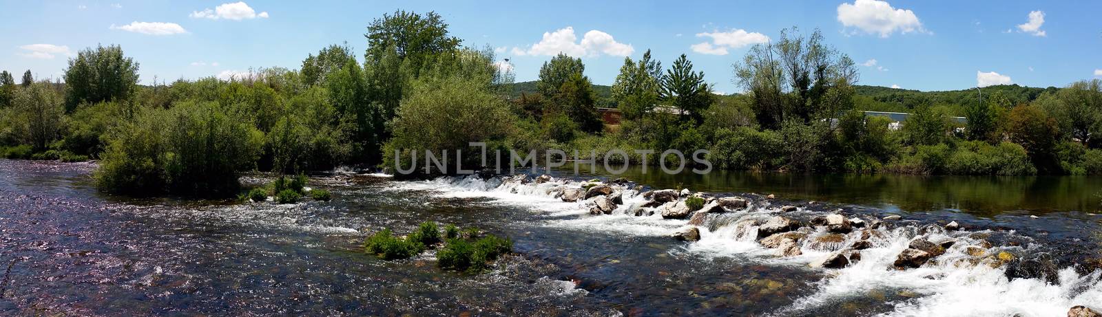 panoramic photo of a waterfall on a small river, with green trees on the opposite side