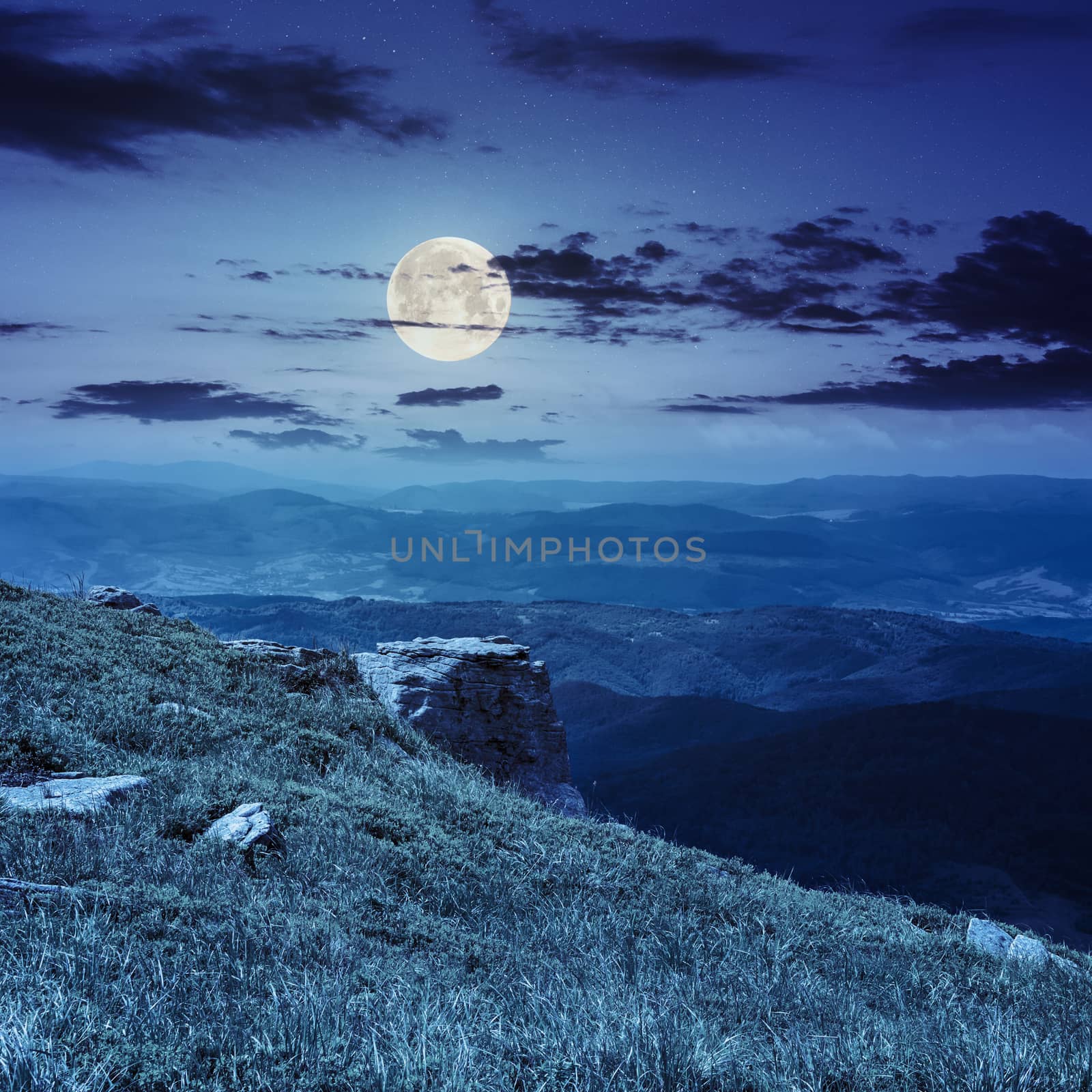 view on high mountains from hillside covered with grass with few stones on the edge at night in full moon light