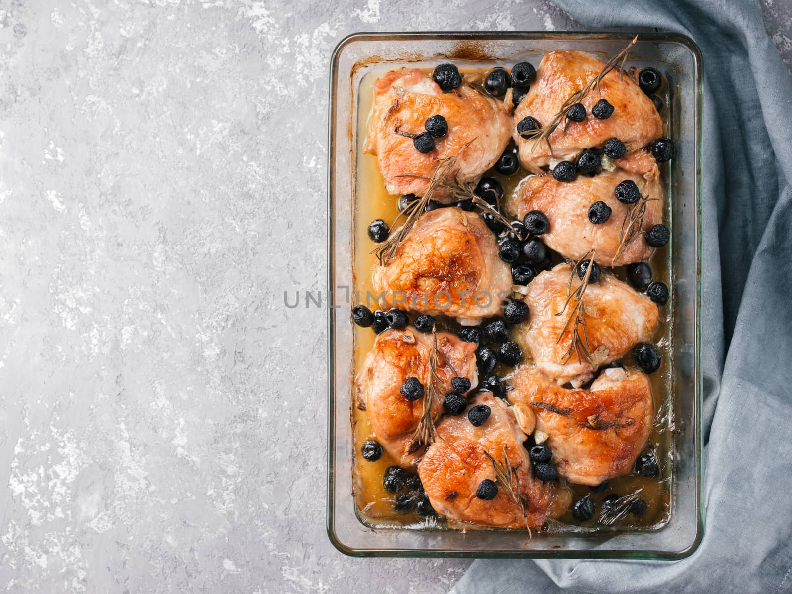 Corsican chicken thighs with rosemary, black olives, garlic in lemon juice and wine. Chicken legs cooked in oven on gray concrete background. Baked chicken leg in heat-proof glass. Top view.Copy space