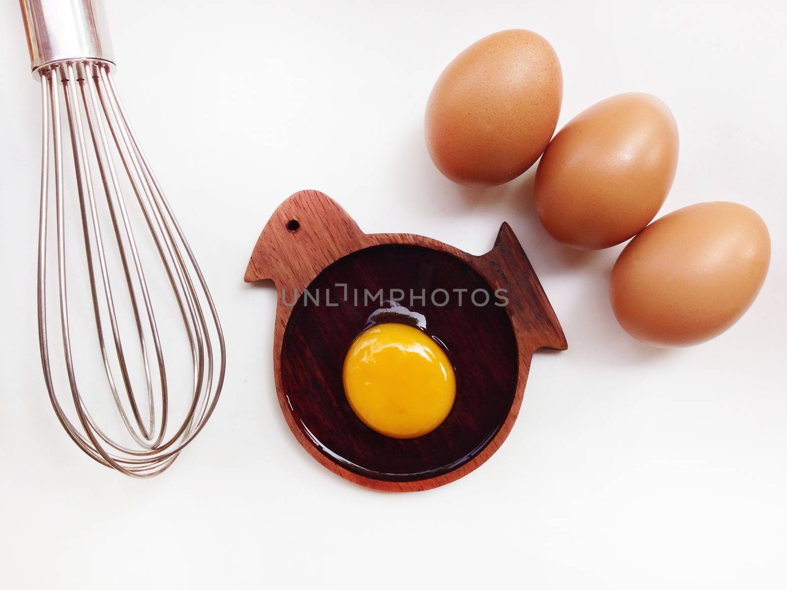 Egg yolk on wooden bird shaped saucer with eggs and egg whisk on white background