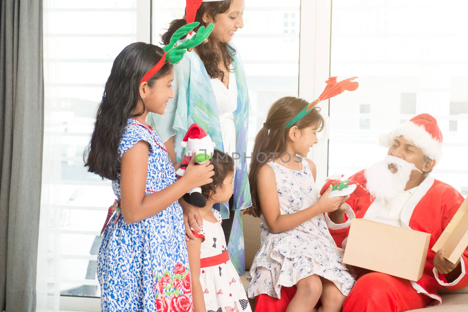 Happy Indian family celebrating Christmas holidays, with gift box and santa hat sitting on sofa or couch at home, Asian parents and children festival mood indoors. 