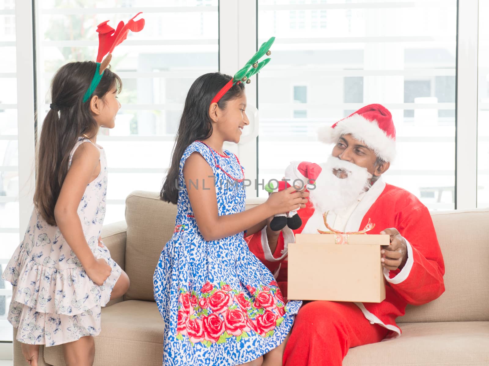 Happy Indian family celebrating Christmas holidays, with gift box and santa sitting on couch at home, Asian people festival mood indoors. 