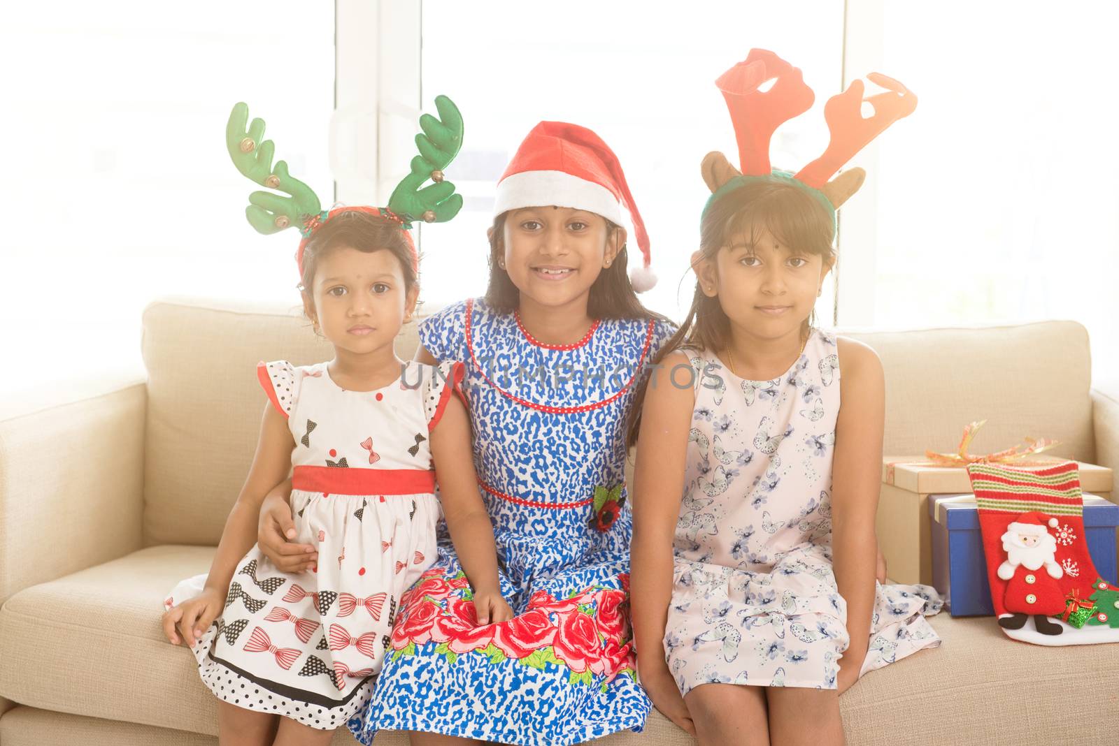 Happy Indian family celebrating Christmas holidays, with gift box and santa hat sitting on sofa or couch at home, adorable Asian children on festival mood. 