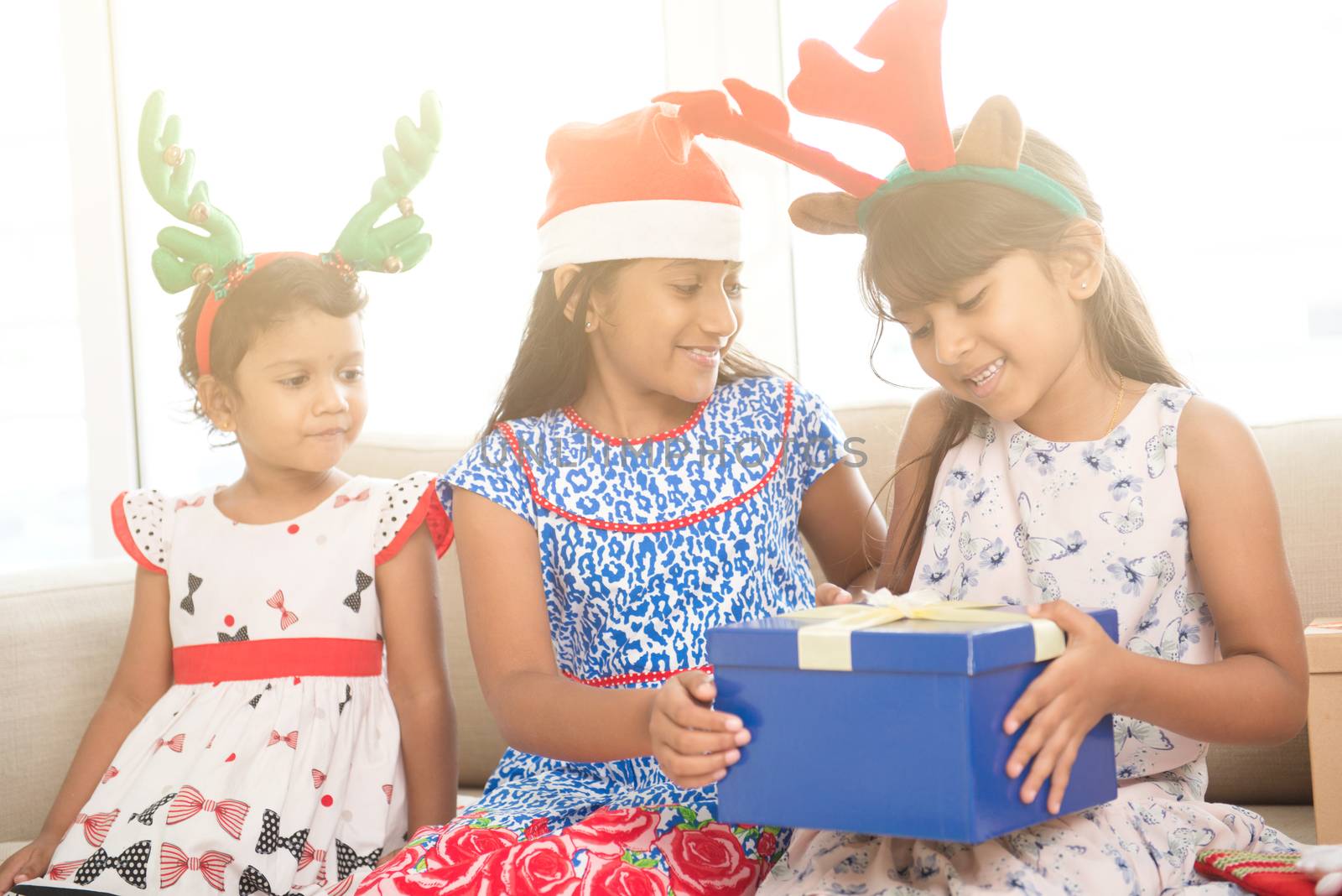 Happy Indian family celebrating Christmas holidays, with gift box and santa hat sitting on couch at home, joyful Asian children on festival mood. 