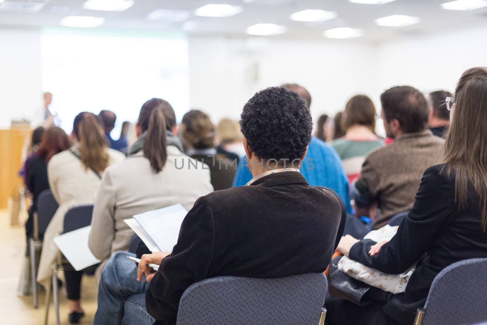 Business and entrepreneurship symposium. Female speaker giving a talk at business meeting. Audience in conference hall. Rear view of unrecognized participants making notes in audience.