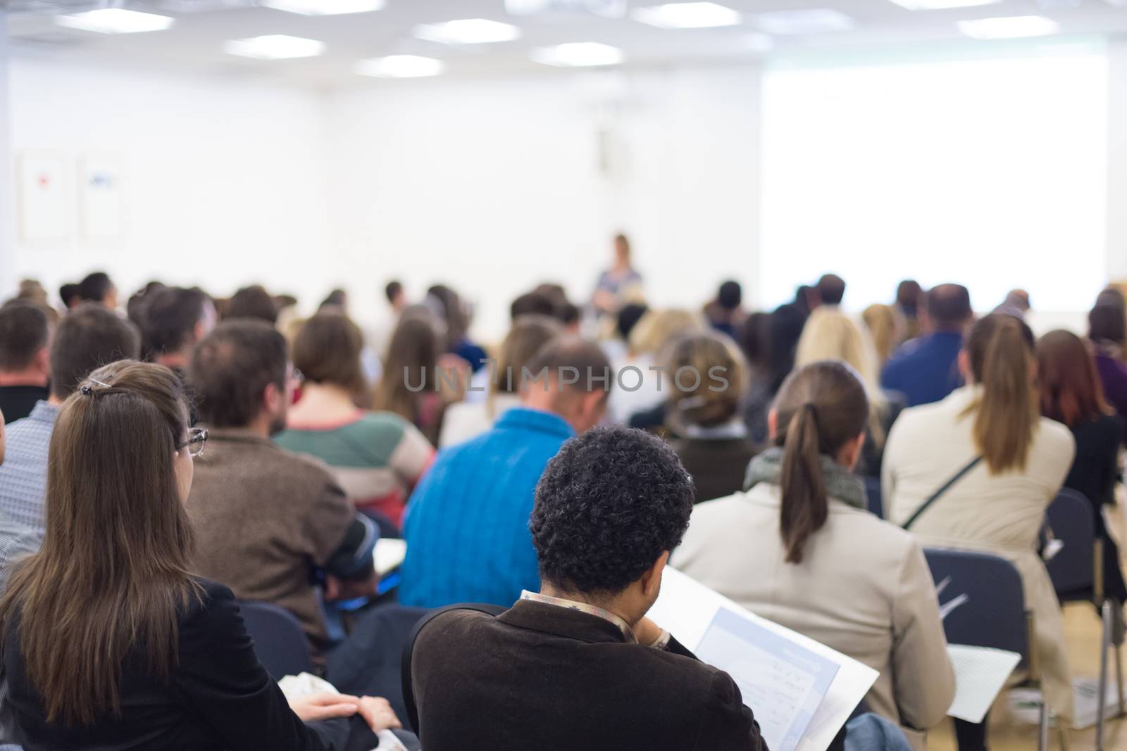 Business and entrepreneurship symposium. Female speaker giving a talk at business meeting. Audience in conference hall. Rear view of unrecognized participants making notes in audience.