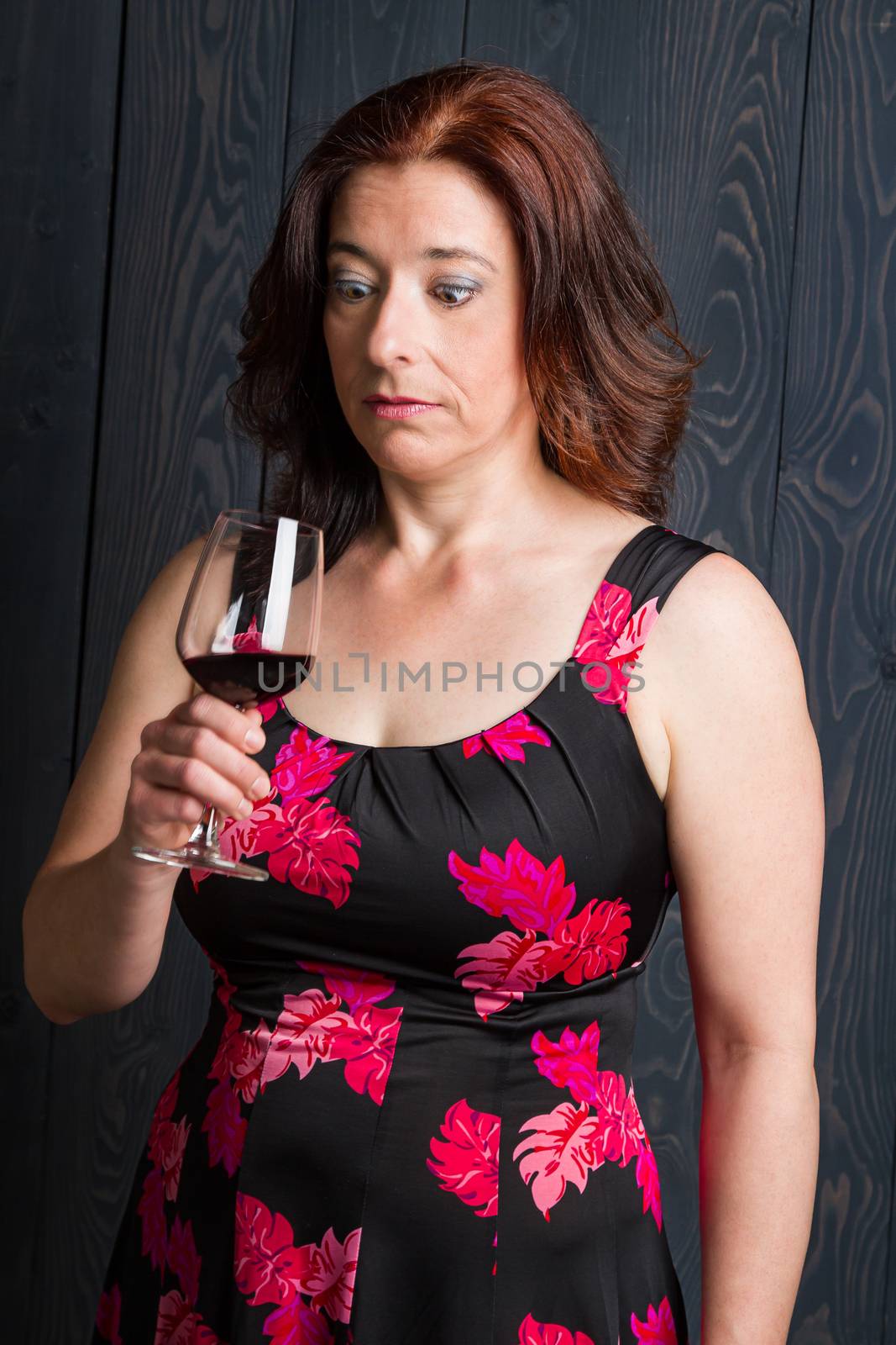 forty something brunette woman wearing a sun dress in front of blue wood panel looking into her wine glass with curious expression