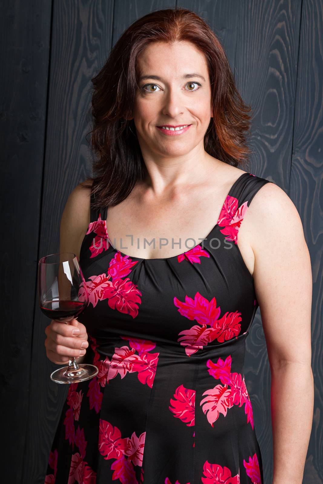 forty something brunette woman wearing a sun dress in front of blue wood panel with a glass of red wine