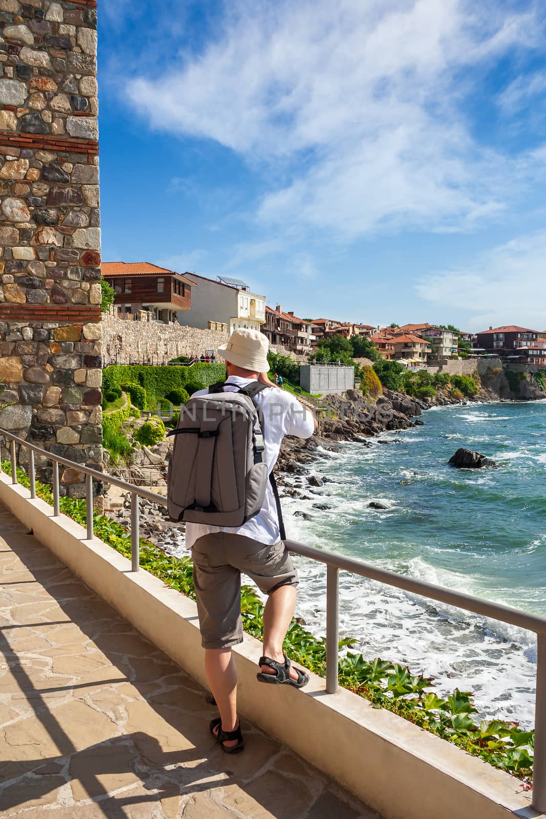 young photographer with backpack  making shots of ancient european town Sozopol on a rocky shore near sea in summer
