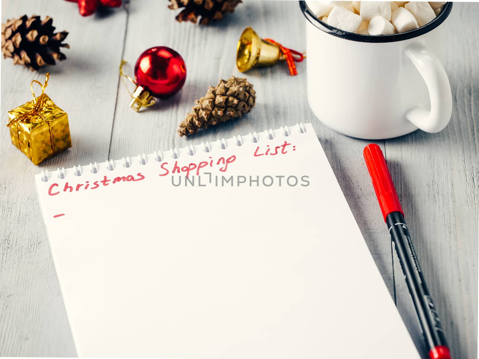 Christmas gifts shopping planning. Make shopping or to-do list for Christmas. Notebook, mug hot chocolate with marshmallows, New Year's decoration and pine cone on gray wooden background.