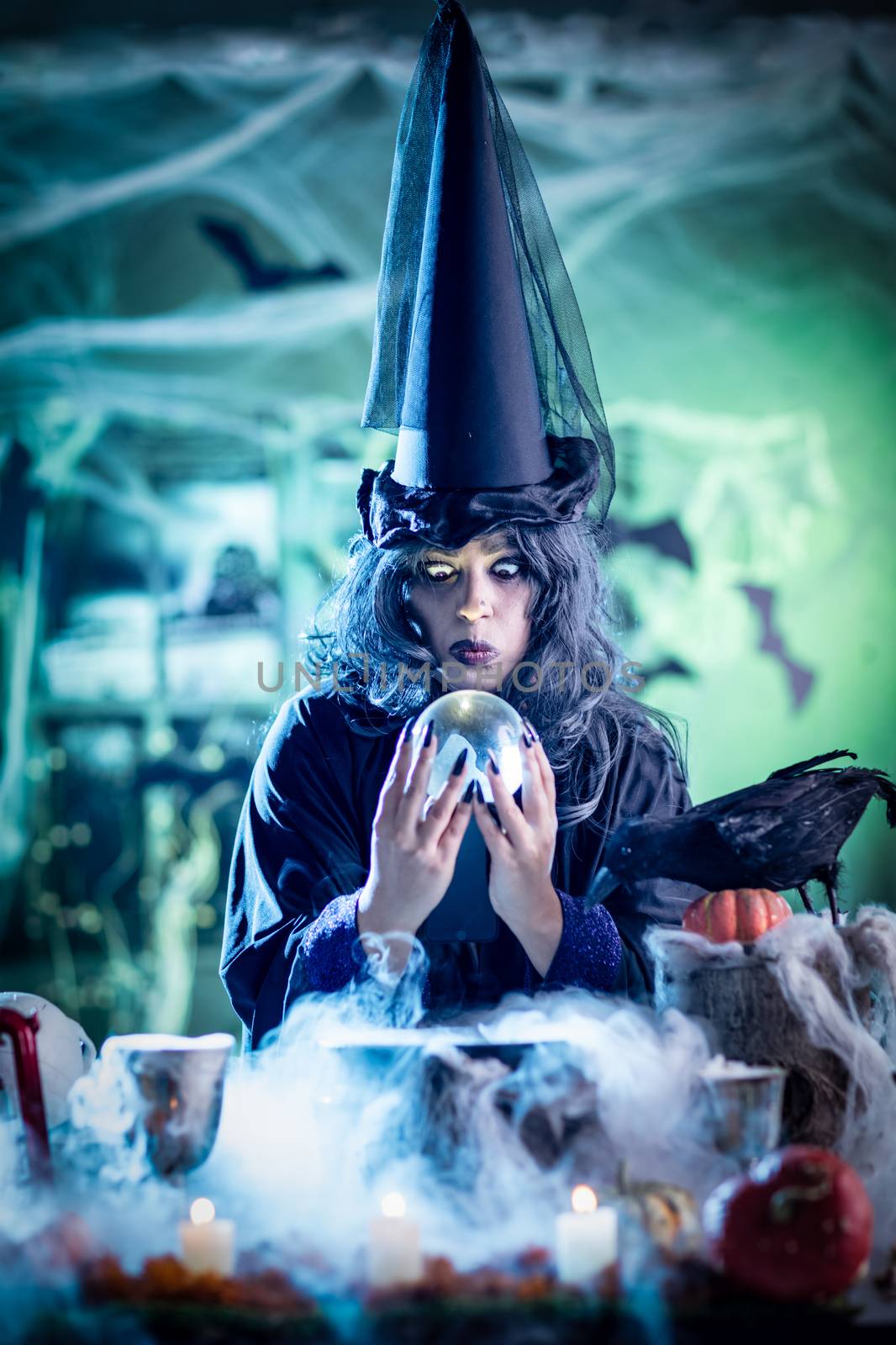 Witch with awfully face in creepy surrounding watching the future in magic ball above boiling potion. 