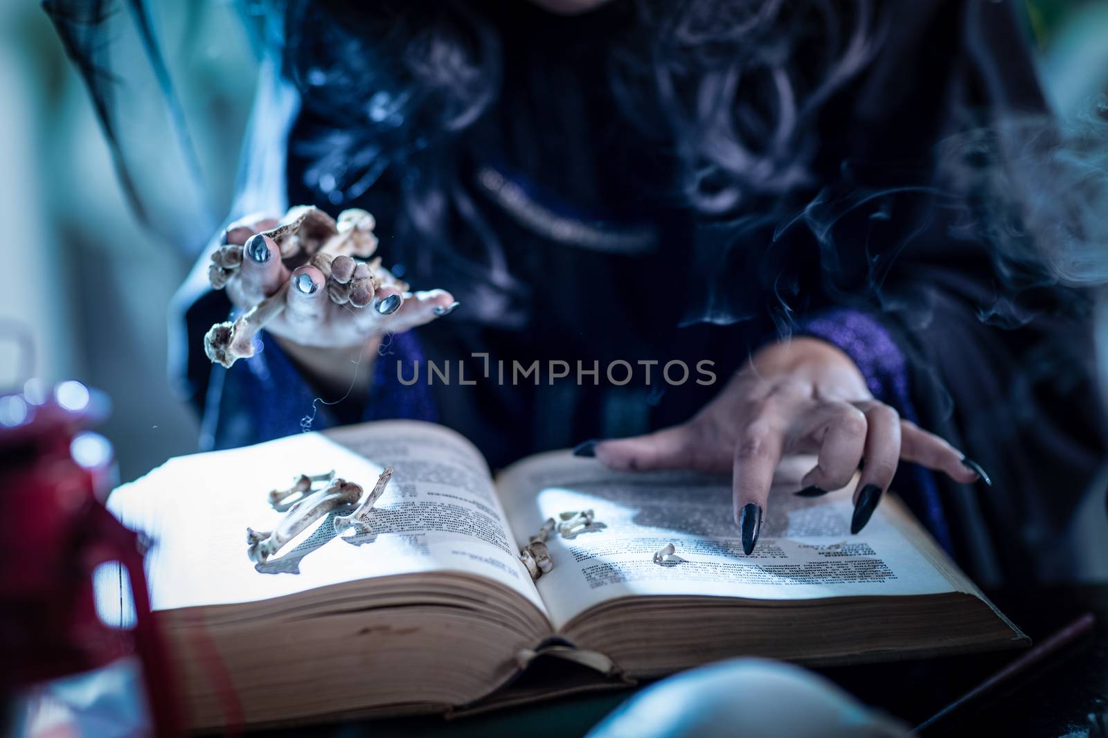 Close up of a witch's hands with pointed black nails holding bones above book of a magic recipe.