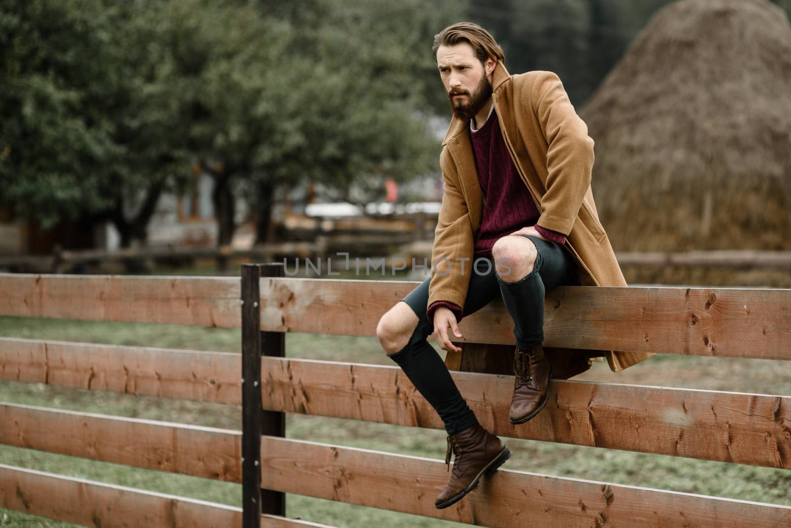 man in a brown coat and torn pants sitting on a wooden fence