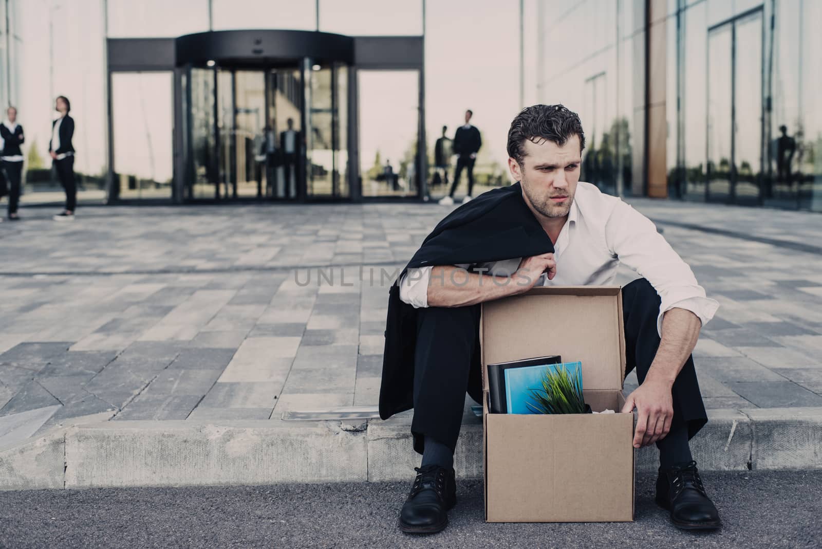 Fired business man sitting frustrated and upset on the street near office building with box of his belongings. He lost work