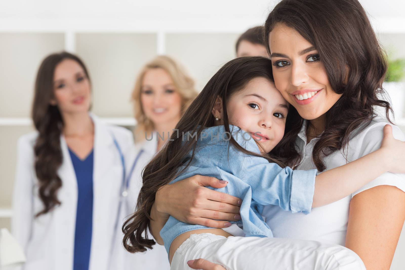 Mother and daughter in medical clinic, team of doctors on background