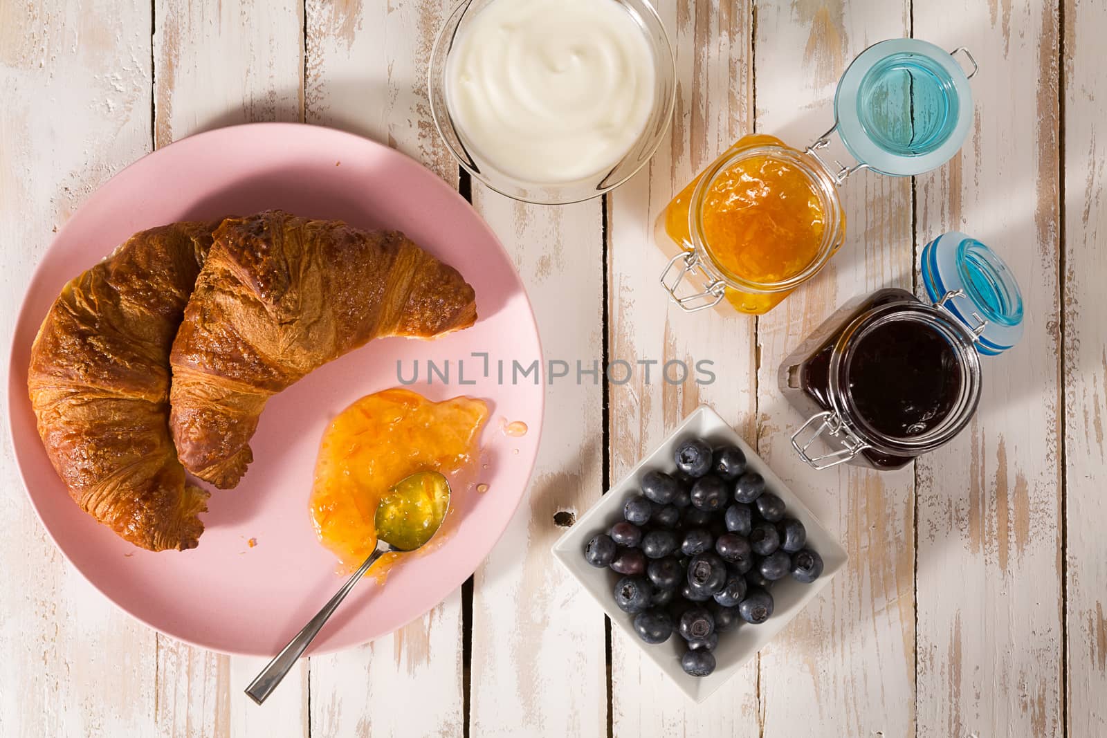 Breakfast with croissant, blueberries, yogurt, orange and blueberry jam over a wooden table seen from above